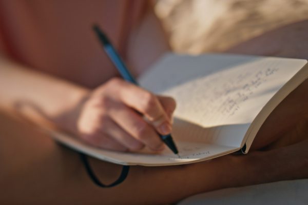 Hands writing personal journal in sunlight closeup. Calm girl rest morning bed noting creative ideas thoughts in diary. Unrecognized woman teenager holding pen making notes. Wish list schedule concept